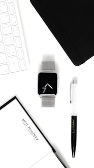 A neatly arranged flat lay featuring a silver smart watch with a mesh band, a black notebook, a clear and black pen with some writing, a white keyboard, and a notebook with a page that has text on it. The items are set against a white background, creating a clean and minimalistic aesthetic.