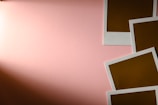 A soft pink background with four blank polaroid-style photo frames arranged in the bottom right corner. The frames have a white border and a brown interior that indicate undeveloped photos.