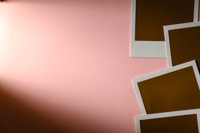 A soft pink background with four blank polaroid-style photo frames arranged in the bottom right corner. The frames have a white border and a brown interior that indicate undeveloped photos.