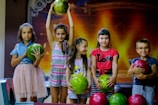 A group of five children is standing indoors, each holding or posing with colorful bowling balls. The background has a warm, glowing pattern that resembles a stage or theater setting. The children appear happy and playful, with one child holding a bowling ball above her head.