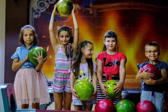 A group of five children is standing indoors, each holding or posing with colorful bowling balls. The background has a warm, glowing pattern that resembles a stage or theater setting. The children appear happy and playful, with one child holding a bowling ball above her head.