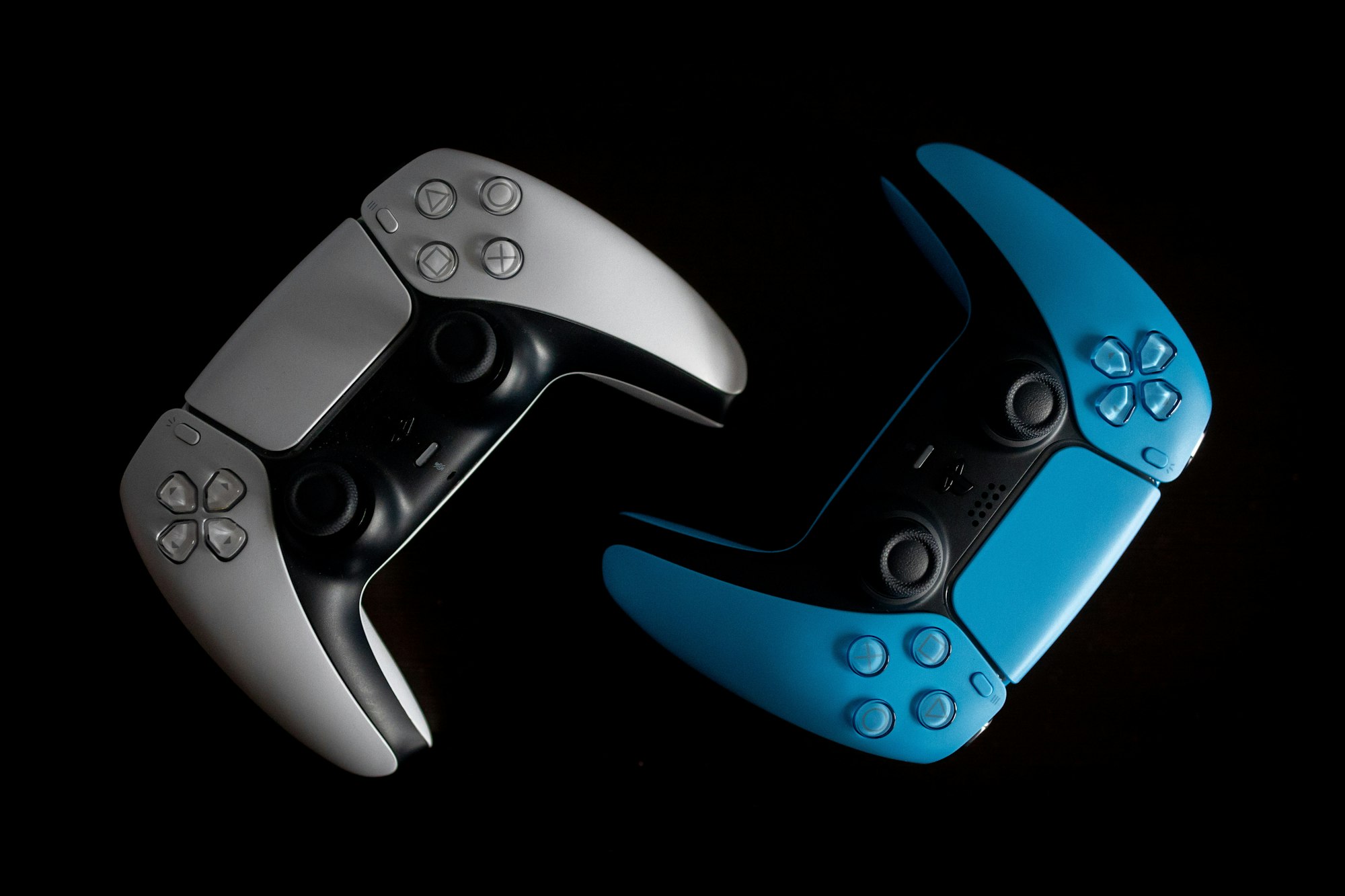 Two PS5 controllers, one white, one teal, on an artful black background.
