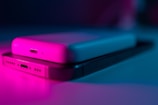A close-up view of a smartphone lying beneath a portable charger. The scene is illuminated with pink and blue lighting, casting a modern and futuristic ambiance over the devices. The focus is on the charging ports and sleek design, emphasizing the connection and functionality of the gadgets.