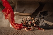 A group of dancers perform a coordinated pose against a modern, geometric concrete backdrop. They are dressed in contrasting outfits with red and black flowing fabric, creating a dynamic sense of movement. The lighting casts strong shadows, enhancing the dramatic effect.