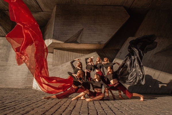 A group of dancers perform a coordinated pose against a modern, geometric concrete backdrop. They are dressed in contrasting outfits with red and black flowing fabric, creating a dynamic sense of movement. The lighting casts strong shadows, enhancing the dramatic effect.