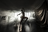 A graceful silhouette of a dancer striking a pose in a dimly lit room filled with fog. The dancer stands on one leg atop a stool, framed by soft lighting from a large window in the background. Various studio equipment, including chairs and a curtain, are partially visible in the shadows.