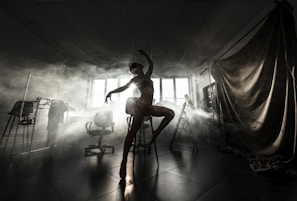 A graceful silhouette of a dancer striking a pose in a dimly lit room filled with fog. The dancer stands on one leg atop a stool, framed by soft lighting from a large window in the background. Various studio equipment, including chairs and a curtain, are partially visible in the shadows.