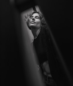 A person with short hair is captured in a moment of serene expression. The black and white photograph creates a dramatic effect with soft lighting and shadows. The background is blurred, drawing focus to the facial features and posture.