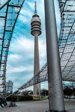 A tall telecommunication tower stands prominently, surrounded by modern architectural elements with a lattice roof structure. The sky is partly cloudy, providing a contrasting backdrop to the tower. In the foreground, there are bicycles parked and sparse trees scattered across a paved area.