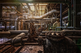 A complex network of industrial pipes and valves with a gritty, aged appearance. The scene includes rusted metal and machinery, suggesting an old factory or mechanical plant. Overhead, the space is dimly lit with light filtering through a grated ceiling, casting shadows and creating an atmospheric setting.