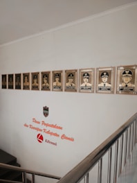 A wall display featuring a series of framed portraits of individuals in uniform, positioned above a staircase. The wall includes text that reads 'Dinas Perpustakaan dan Kearsipan Kabupaten Ciamis Literasi,' accompanied by a logo shield.