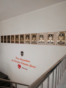 A wall display featuring a series of framed portraits of individuals in uniform, positioned above a staircase. The wall includes text that reads 'Dinas Perpustakaan dan Kearsipan Kabupaten Ciamis Literasi,' accompanied by a logo shield.