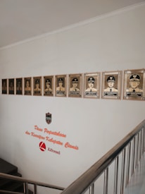 A wall display featuring a series of framed portraits of individuals in uniform, positioned above a staircase. The wall includes text that reads 'Dinas Perpustakaan dan Kearsipan Kabupaten Ciamis Literasi,' accompanied by a logo shield.
