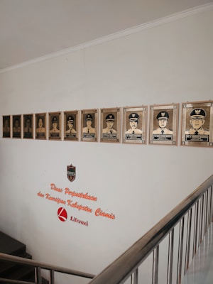 A wall display featuring a series of framed portraits of individuals in uniform, positioned above a staircase. The wall includes text that reads 'Dinas Perpustakaan dan Kearsipan Kabupaten Ciamis Literasi,' accompanied by a logo shield.
