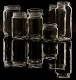 Several empty glass jars are arranged in a group on a reflective black surface, creating a clear reflection beneath. The lighting casts highlights on the glass, showcasing their transparency and shape.