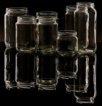 Several empty glass jars are arranged in a group on a reflective black surface, creating a clear reflection beneath. The lighting casts highlights on the glass, showcasing their transparency and shape.