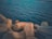 Large, geometric concrete blocks are situated by the edge of a calm body of water. The water's surface is rippled with gentle waves, extending to the horizon. The light casts a warm tone on the concrete structures, creating a contrast with the deep blue of the sea.