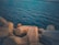Large, geometric concrete blocks are situated by the edge of a calm body of water. The water's surface is rippled with gentle waves, extending to the horizon. The light casts a warm tone on the concrete structures, creating a contrast with the deep blue of the sea.