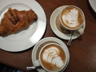 A warm brown wooden table with a freshly baked croissant on a white plate, accompanied by two cups of cappuccino featuring intricate leaf latte art.