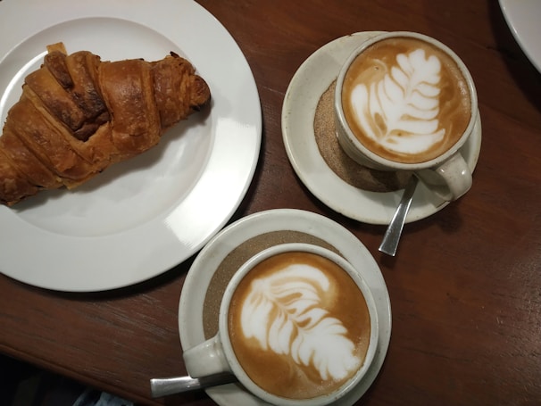 A rustic wooden table with a steaming cup of artisan coffee and a freshly baked croissant beside it.