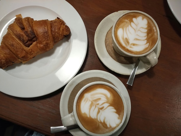 A warm brown wooden table with a freshly baked croissant on a white plate, accompanied by two cups of cappuccino featuring intricate leaf latte art.