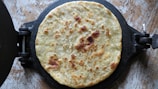 A freshly made roti or flatbread is resting on a black roti maker with visible browned spots, indicating it has been cooked. The background surface appears to be a textured, light-colored stone or wood.