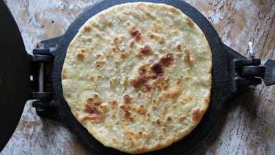 A freshly made roti or flatbread is resting on a black roti maker with visible browned spots, indicating it has been cooked. The background surface appears to be a textured, light-colored stone or wood.