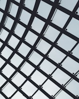 A geometric grid structure made up of intersecting metal beams and mesh panels against a light sky background. The pattern forms a series of rectangles with uniform spacing, conveying a sense of order and modern architectural design.