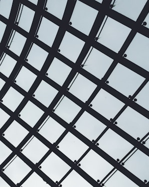 A geometric grid structure made up of intersecting metal beams and mesh panels against a light sky background. The pattern forms a series of rectangles with uniform spacing, conveying a sense of order and modern architectural design.