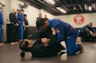 A group of people are practicing Brazilian Jiu-Jitsu in a martial arts gym. Two individuals are actively engaged in grappling on the mats while others observe or participate in the background. Many are wearing blue and black gis, typical attire for this type of martial art. The atmosphere appears focused and disciplined, with a sense of concentration on the training.