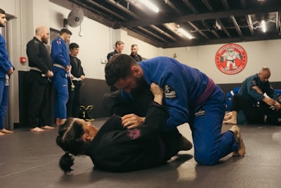 A group of people are practicing Brazilian Jiu-Jitsu in a martial arts gym. Two individuals are actively engaged in grappling on the mats while others observe or participate in the background. Many are wearing blue and black gis, typical attire for this type of martial art. The atmosphere appears focused and disciplined, with a sense of concentration on the training.
