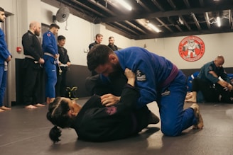 A group of people are practicing Brazilian Jiu-Jitsu in a martial arts gym. Two individuals are actively engaged in grappling on the mats while others observe or participate in the background. Many are wearing blue and black gis, typical attire for this type of martial art. The atmosphere appears focused and disciplined, with a sense of concentration on the training.