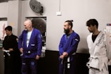 Four individuals stand in a martial arts studio, wearing Brazilian jiu-jitsu gis in various colors including black, blue, and white. They hold belts and appear attentive, possibly awaiting instruction or recognition.