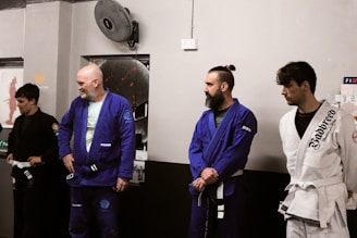 Four individuals stand in a martial arts studio, wearing Brazilian jiu-jitsu gis in various colors including black, blue, and white. They hold belts and appear attentive, possibly awaiting instruction or recognition.