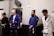 Four individuals stand in a martial arts studio, wearing Brazilian jiu-jitsu gis in various colors including black, blue, and white. They hold belts and appear attentive, possibly awaiting instruction or recognition.