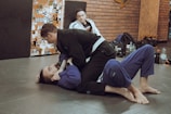 Two individuals are practicing Brazilian Jiu-Jitsu on a mat, both wearing gi uniforms. One person is on the ground while the other is on top, engaging in a grappling move. A third person in a gi is sitting in the background, observing the activity. The environment includes a wall covered with various stickers and gym equipment like bags and bottles are placed around.