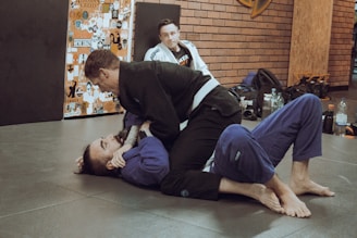 Two individuals are practicing Brazilian Jiu-Jitsu on a mat, both wearing gi uniforms. One person is on the ground while the other is on top, engaging in a grappling move. A third person in a gi is sitting in the background, observing the activity. The environment includes a wall covered with various stickers and gym equipment like bags and bottles are placed around.