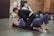 Two individuals are practicing Brazilian Jiu-Jitsu on a mat, both wearing gi uniforms. One person is on the ground while the other is on top, engaging in a grappling move. A third person in a gi is sitting in the background, observing the activity. The environment includes a wall covered with various stickers and gym equipment like bags and bottles are placed around.