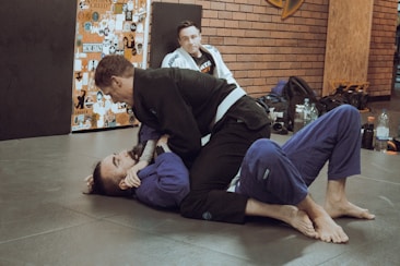 Two individuals are practicing Brazilian Jiu-Jitsu on a mat, both wearing gi uniforms. One person is on the ground while the other is on top, engaging in a grappling move. A third person in a gi is sitting in the background, observing the activity. The environment includes a wall covered with various stickers and gym equipment like bags and bottles are placed around.