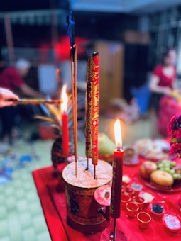 A vibrant and colorful setting features red incense sticks, some burning with small flames, placed in a decorated holder. This is situated on a red cloth, surrounded by various small bowls and a spread of food, including fruits and a pineapple. In the background, a blurred figure can be seen, and the atmosphere suggests a ritual or celebration.