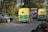 A busy road scene featuring two auto rickshaws painted in yellow and green, driving alongside several other vehicles including a car. The surrounding area is lush with greenery and trees, indicating a rural or semi-urban setting.