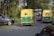 A busy road scene featuring two auto rickshaws painted in yellow and green, driving alongside several other vehicles including a car. The surrounding area is lush with greenery and trees, indicating a rural or semi-urban setting.