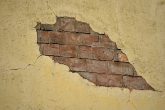 A section of a wall with cracked plaster revealing red bricks underneath. The plaster appears worn and aged, showcasing textures and shades of yellow. The exposed bricks are arranged in a regular pattern, highlighting the contrast between the rough surface of the plaster and the structured brickwork.
