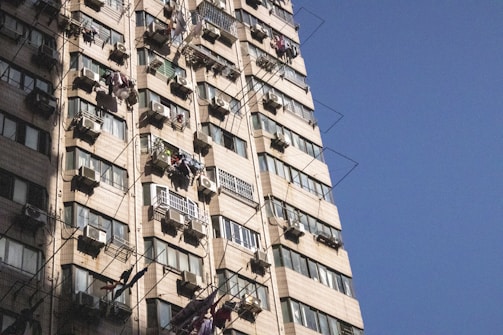 A tall residential building with multiple air conditioning units and laundry hanging from many windows. The building facade features beige tiles and a pattern of windows with metal bars. Shadows are cast on the building, suggesting a sunny day with clear blue sky.