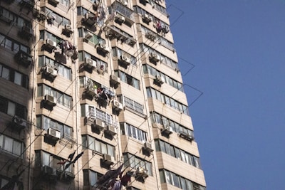 A tall residential building with multiple air conditioning units and laundry hanging from many windows. The building facade features beige tiles and a pattern of windows with metal bars. Shadows are cast on the building, suggesting a sunny day with clear blue sky.