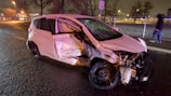 A severely damaged white car is parked on the side of a wet road at night. The front and side of the vehicle are significantly crumpled, and debris is scattered around it. A speed limit sign is visible in the background, along with a sidewalk and fence. There is a person in a hooded jacket walking on the sidewalk.