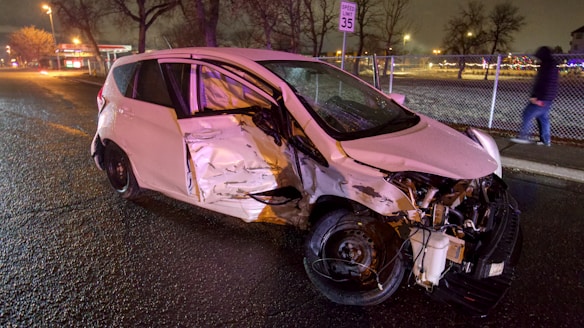 A severely damaged white car is parked on the side of a wet road at night. The front and side of the vehicle are significantly crumpled, and debris is scattered around it. A speed limit sign is visible in the background, along with a sidewalk and fence. There is a person in a hooded jacket walking on the sidewalk.