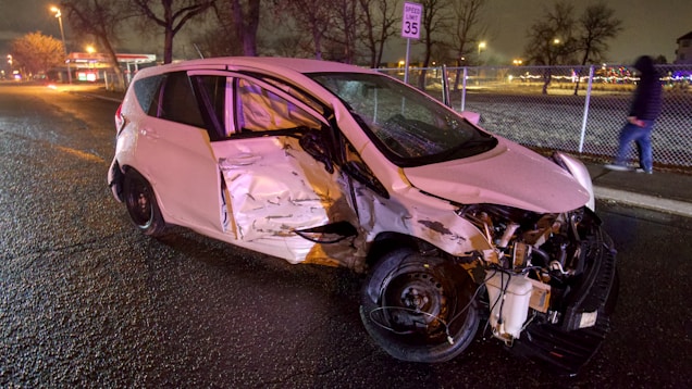 A severely damaged white car is parked on the side of a wet road at night. The front and side of the vehicle are significantly crumpled, and debris is scattered around it. A speed limit sign is visible in the background, along with a sidewalk and fence. There is a person in a hooded jacket walking on the sidewalk.