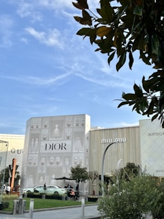 A high-end shopping area features prominently branded stores such as Dior, Gucci, Miu Miu, and Bottega Veneta. The exterior of the Dior store is adorned with artistic depictions of dresses and fashion items. Several people are sitting and walking in the outdoor space, surrounded by well-maintained greenery and trees. The sky is clear with a few clouds, adding to the relaxed atmosphere of the scene.
