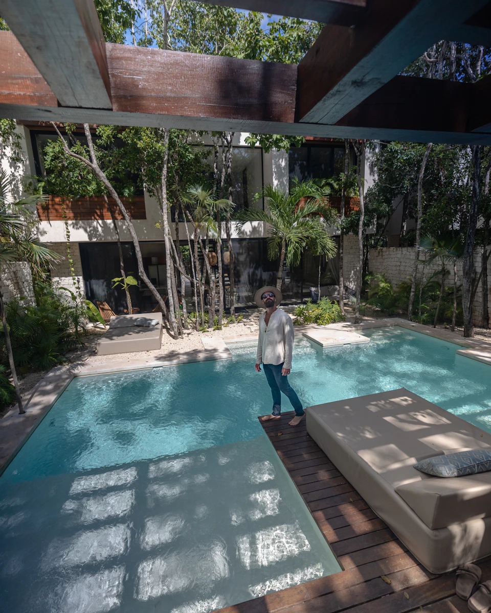 The image depicts a serene outdoor setting with a large, rectangular swimming pool filled with clear blue water and surrounded by lush greenery. A woman is seen walking towards the pool area, adding a sense of scale to the scene. The pool's edge features a wooden deck that extends into the distance, providing a natural transition between the pool and the surrounding environment. The image is taken from an elevated perspective, offering a comprehensive view of the pool and its surroundings.